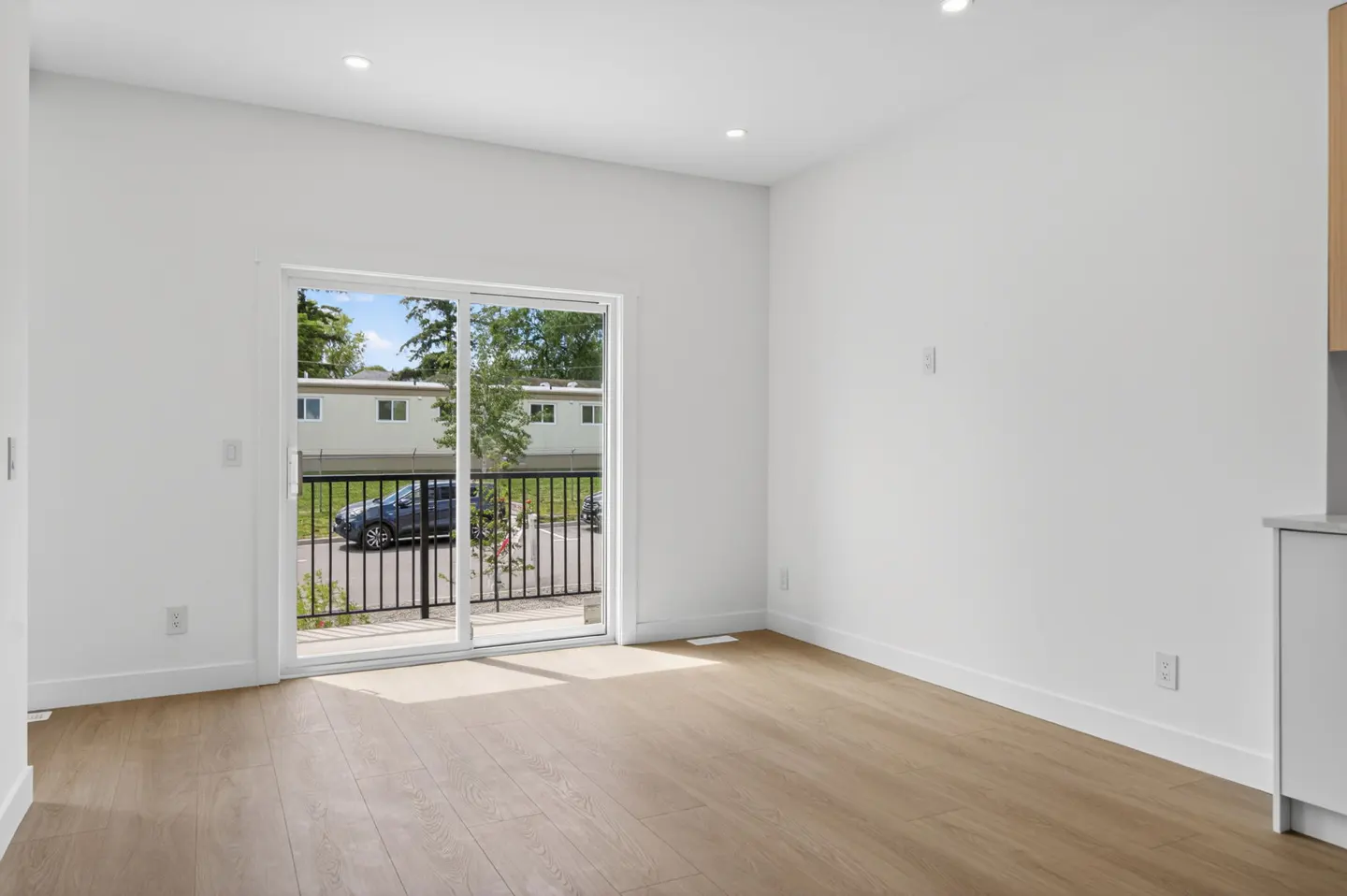 Bright, empty room with white walls, light wood floors, and a sliding glass door leading to a balcony with a parking lot.