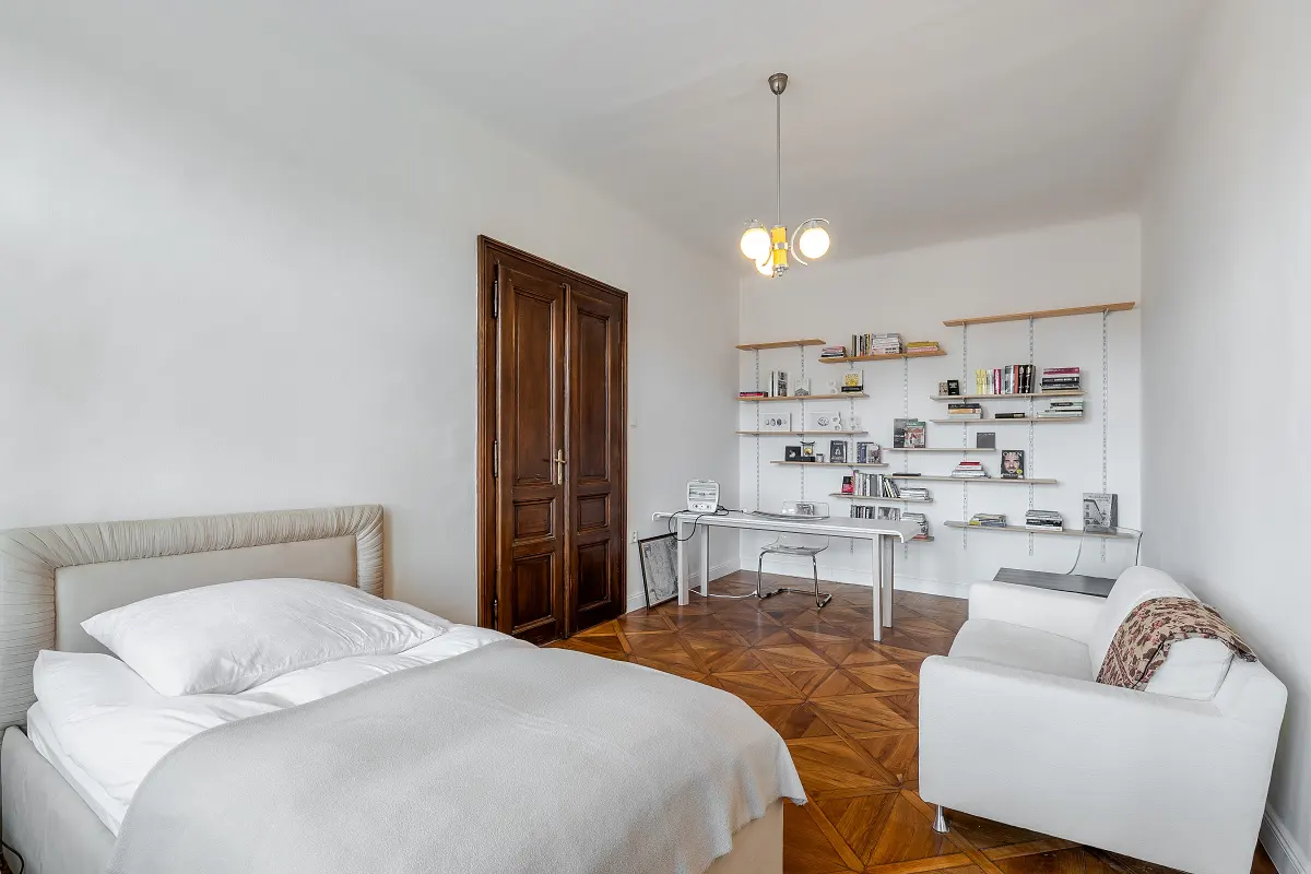 Bright bedroom with a bed, white sofa, desk with shelves, and parquet floor.