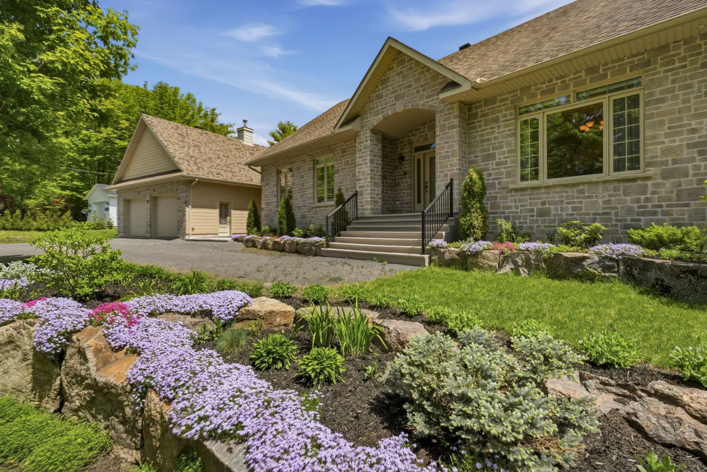 Stone house with a detached garage, front steps, and a landscaped garden featuring purple flowers and rock retaining walls.