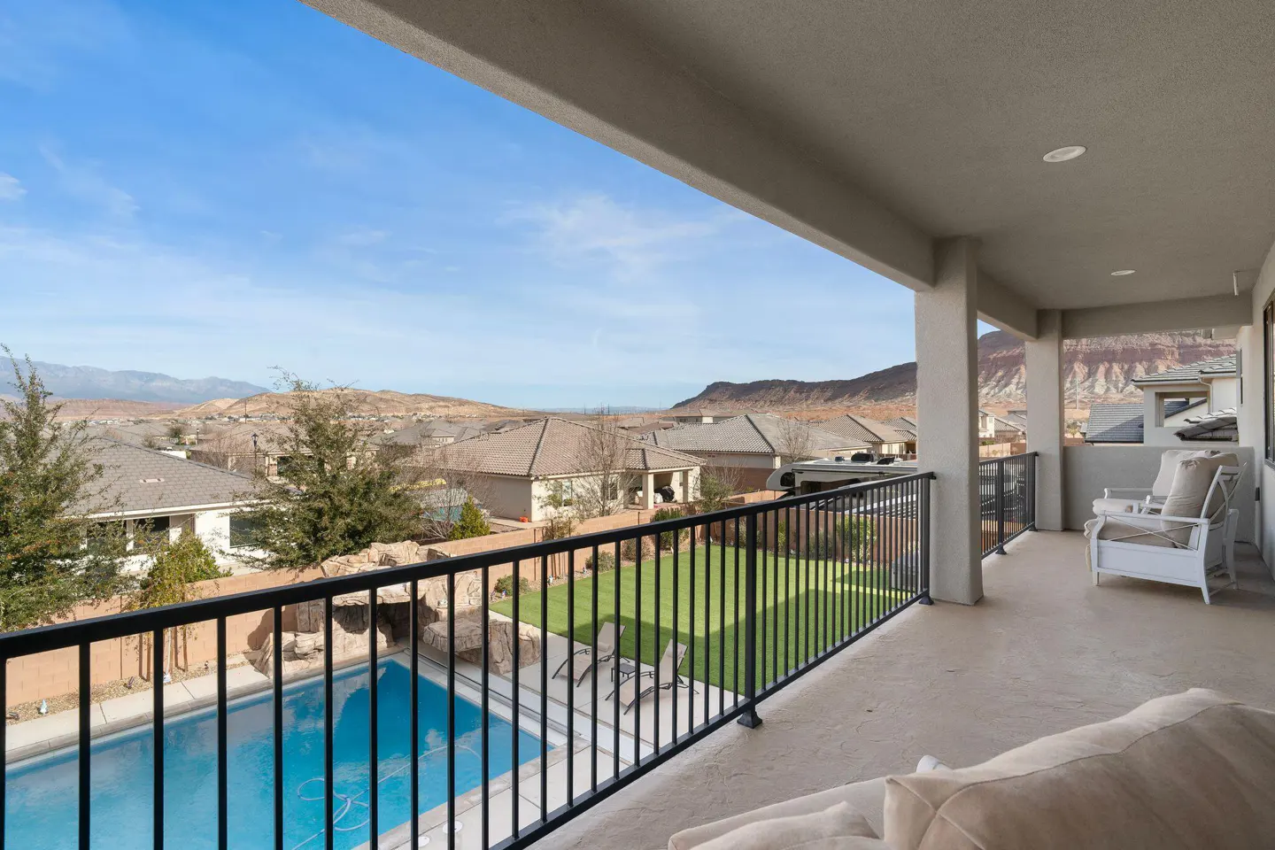 Balcony view of a pool, lawn, and houses under a blue sky. Black metal railing and white patio furniture are visible.