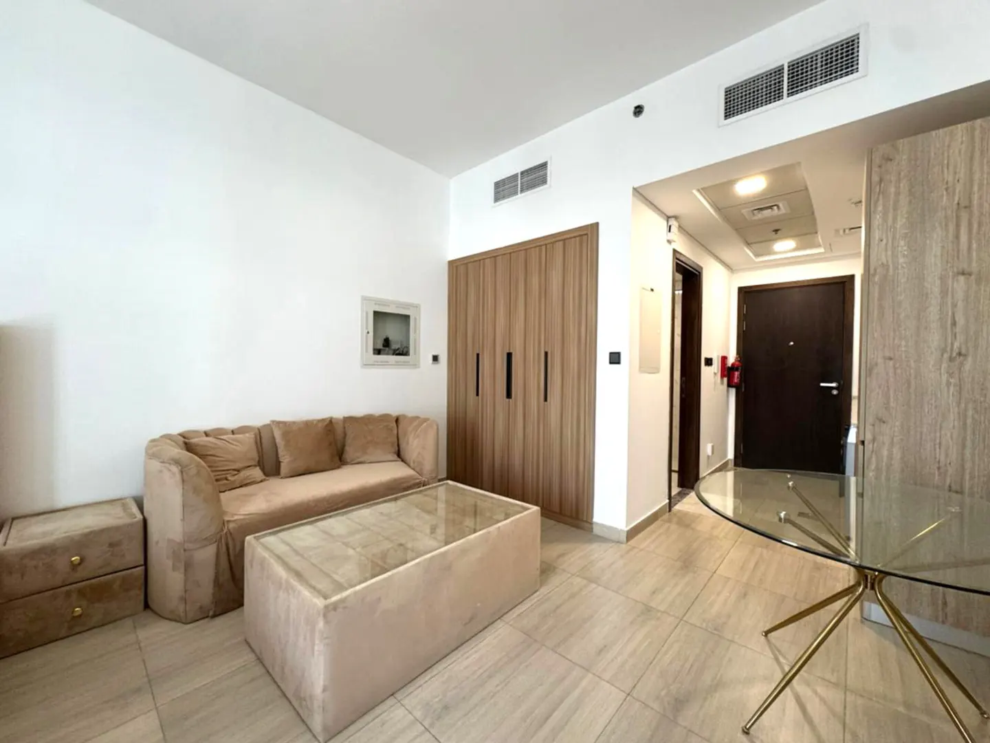A bright living room with a beige sofa, glass coffee table, and wood-look cabinets. A round glass dining table sits near the doorway.