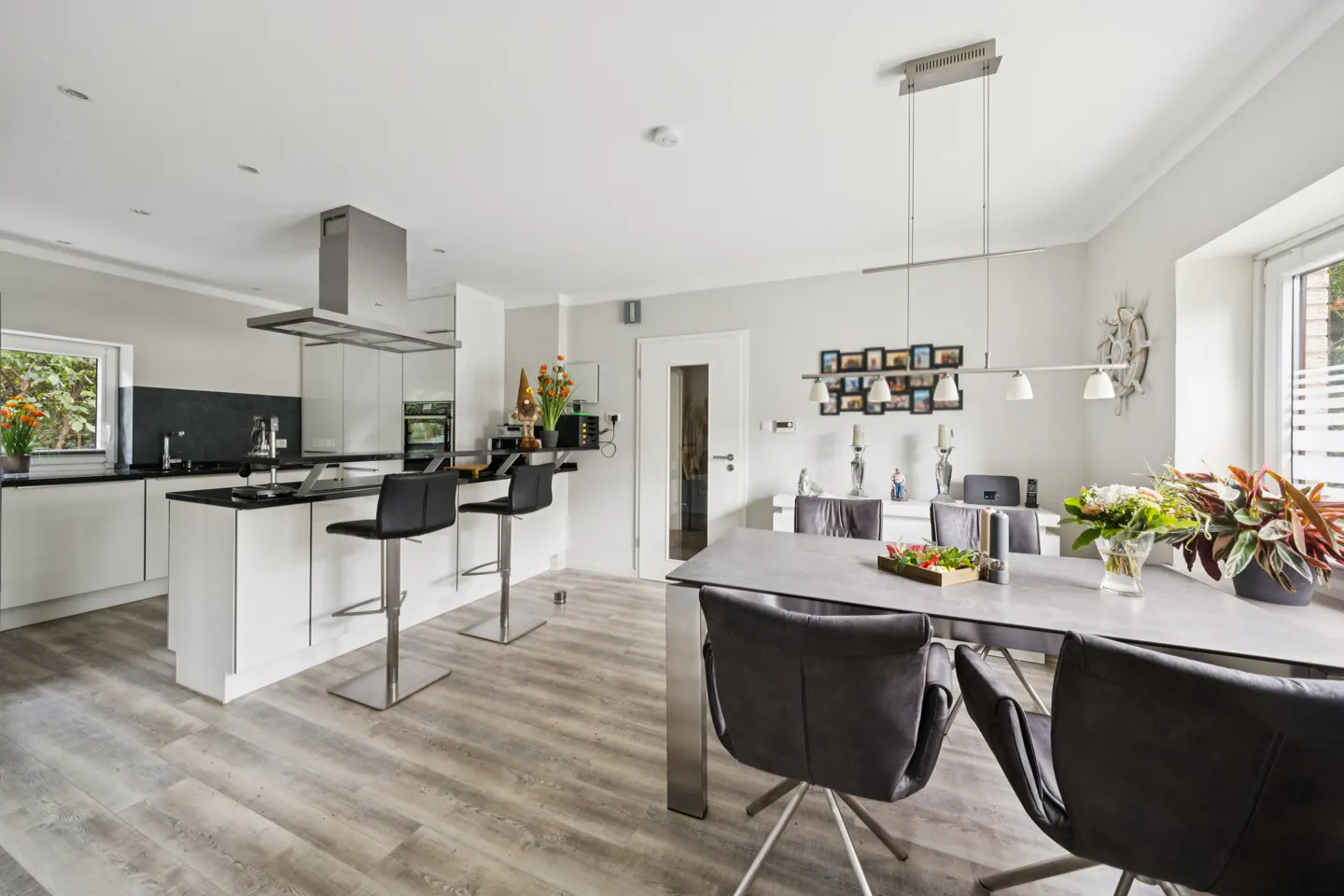 Bright, modern kitchen and dining area with white cabinets, black countertops, and gray wood-look floors. A dining table with gray chairs is visible.