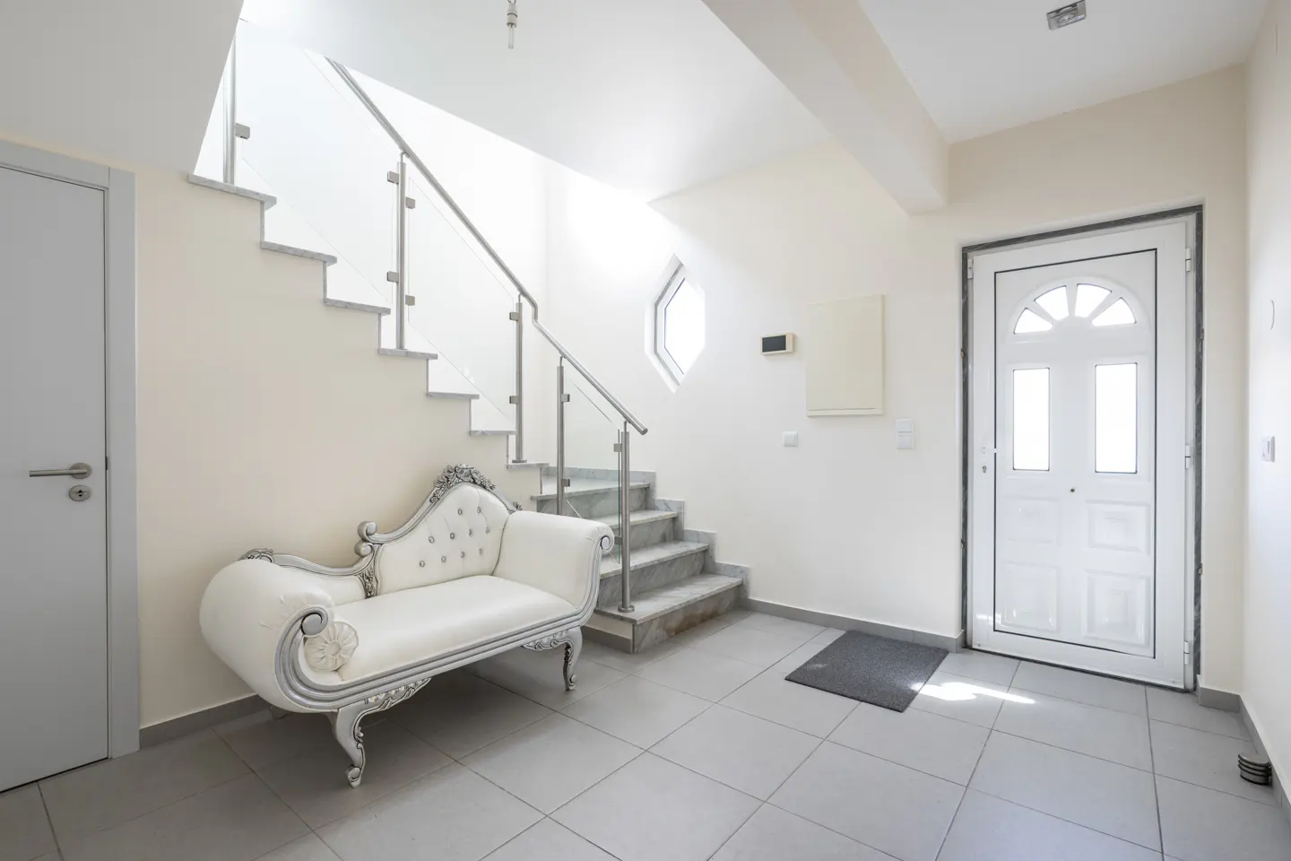 Bright foyer with gray tile floor, white walls, and staircase. A white chaise lounge sits under the stairs, near a white front door.