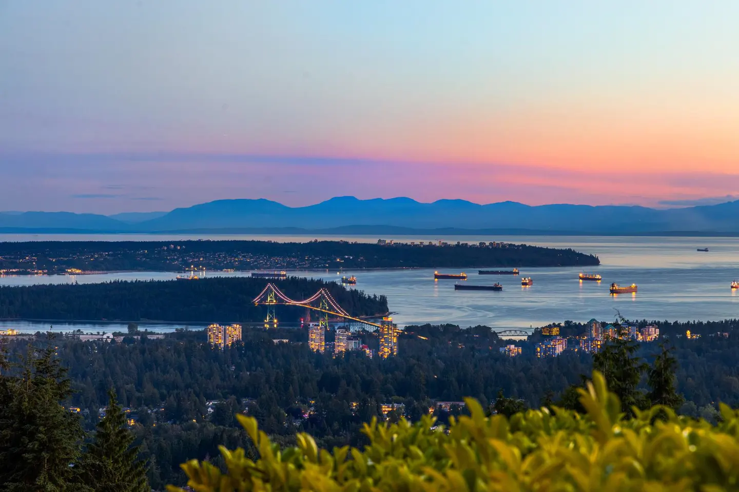 Vancouver skyline at dusk. The Lions Gate Bridge spans the water, with cargo ships visible. Mountains are in the background.