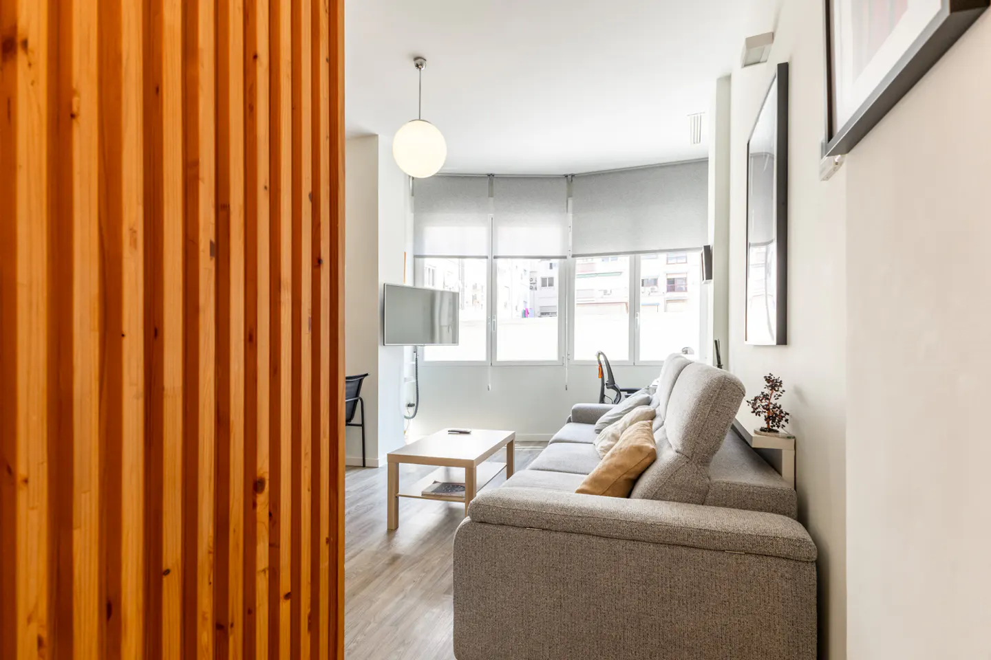 Living room with a gray sofa, wood floors, and a light wood slat wall. Windows with white blinds let in natural light.