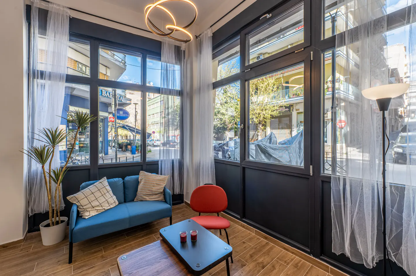 Bright room with large windows showing a city street. A blue loveseat, red chair, and coffee table furnish the space. Sheer white curtains soften the light.