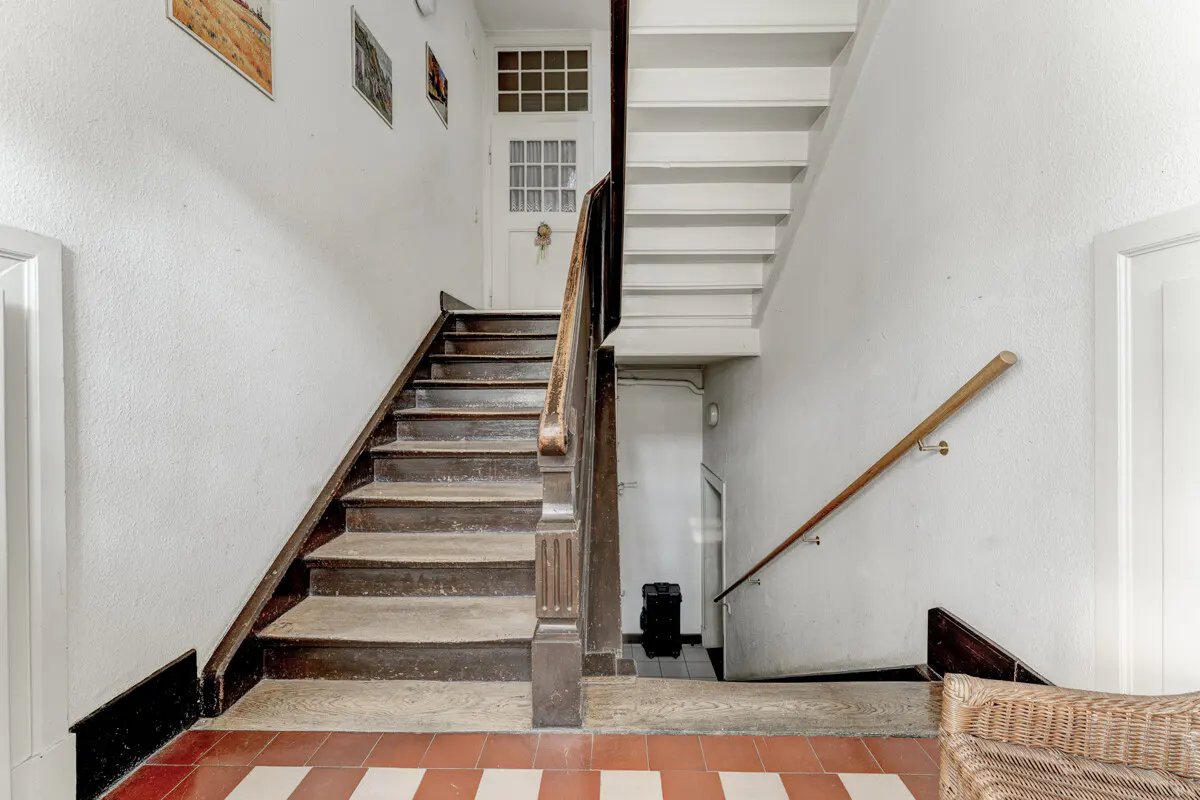 Interior view of a white stairwell with wooden stairs and handrails, red and white tiled floor, and artwork on the walls.