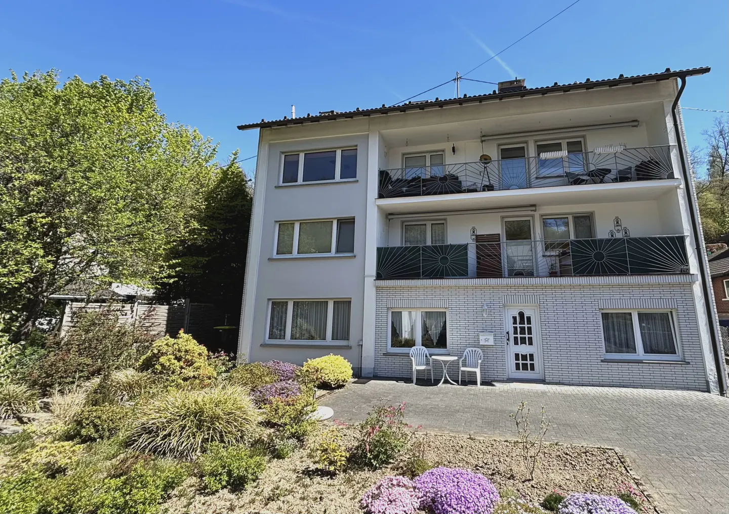 Two-story white building with balconies and a white brick facade. A small table and chairs sit outside the front door. Landscaped yard with colorful flowers.