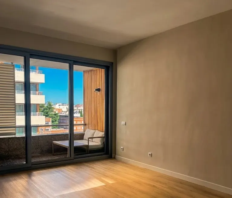 Empty room with wood floors and beige walls. Sliding glass doors open to a balcony with a couch and city view.
