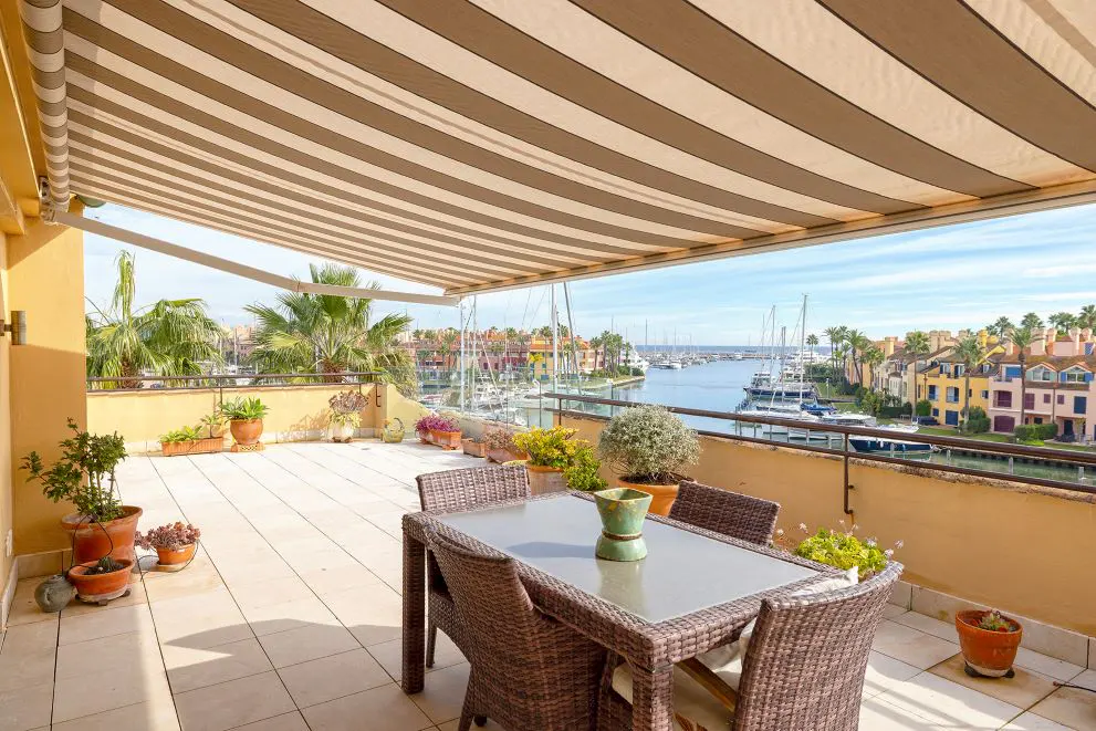 Outdoor patio with a table and chairs overlooking a marina with boats and colorful buildings. A striped awning provides shade.