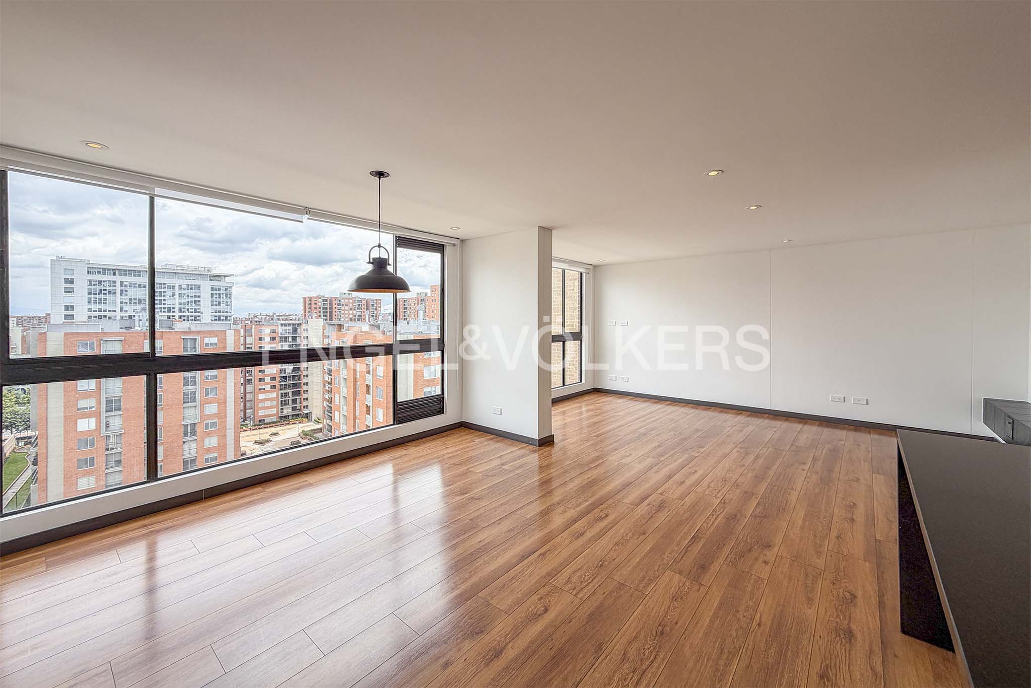 Bright, empty living room with wood floors, white walls, and large windows overlooking a city. A black pendant light hangs from the ceiling.