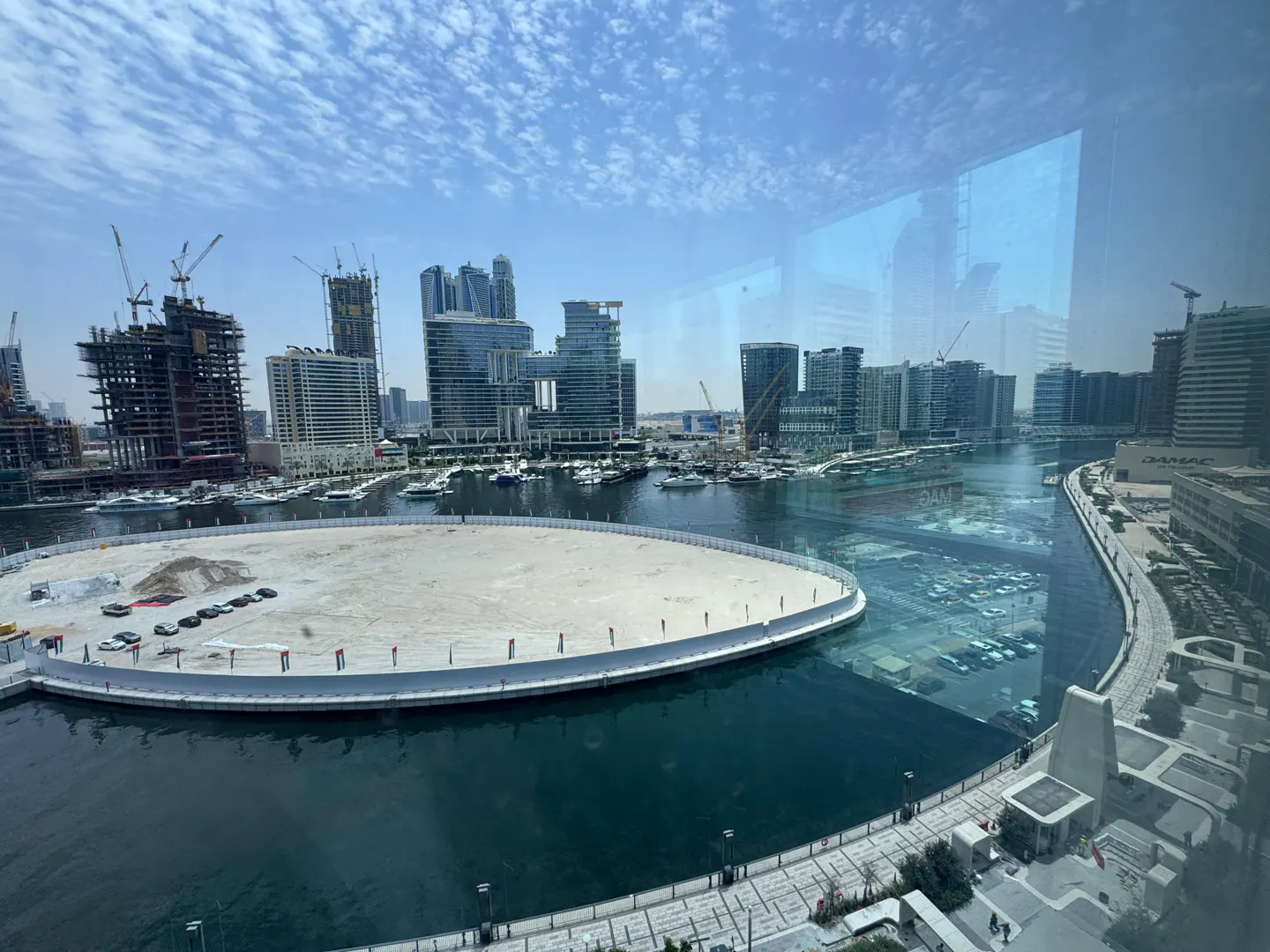 View of a city skyline with modern buildings, cranes, and a waterway with boats, under a blue sky.
