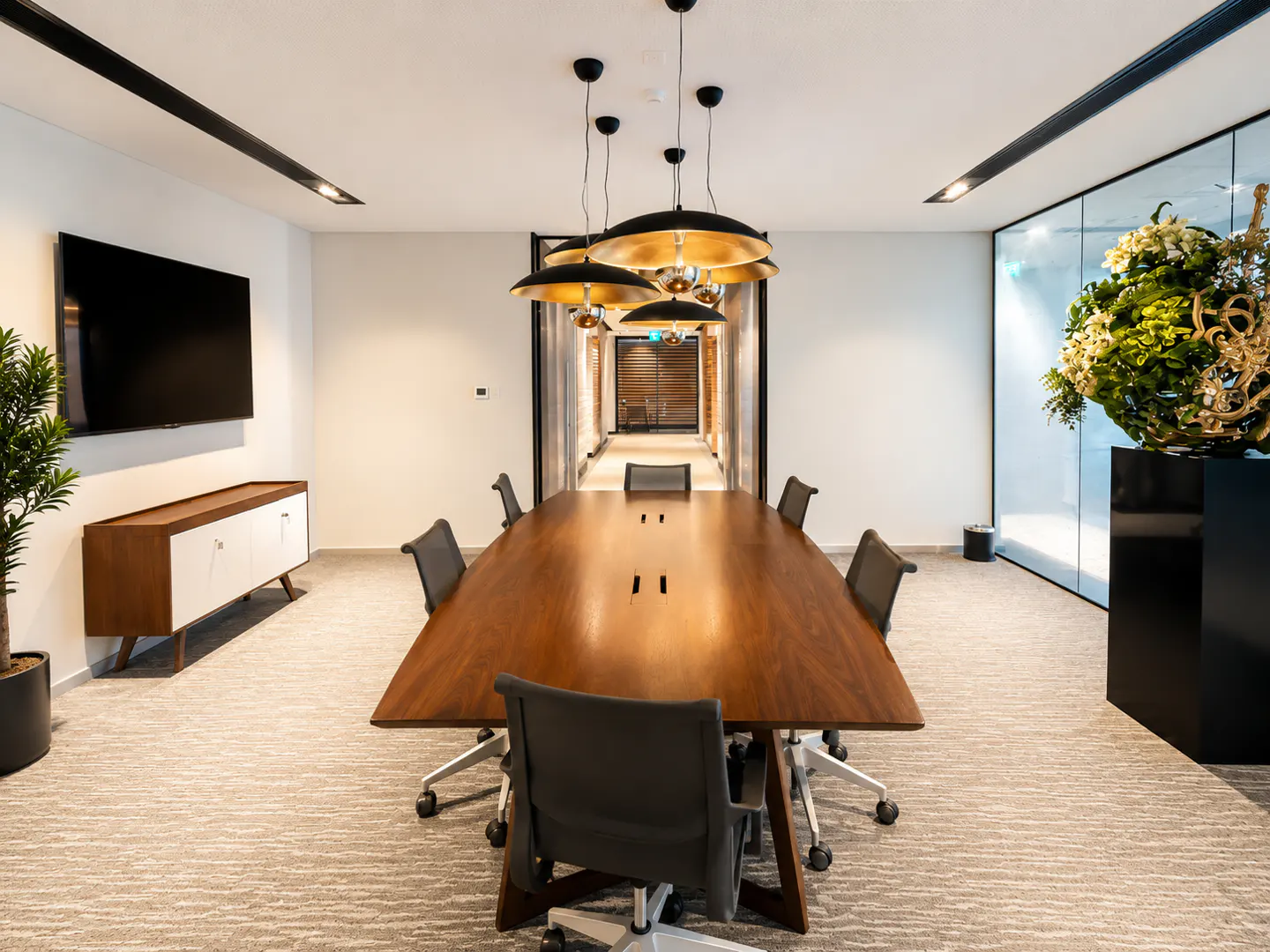 Modern boardroom with a long wooden table, black chairs, hanging pendant lights, a wall-mounted TV, and floral decor.