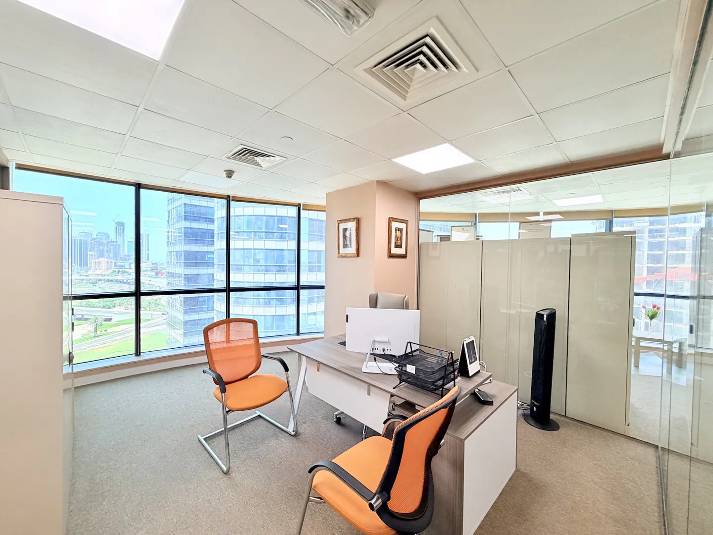 Bright office space with two orange chairs, a desk with a computer, and a large window overlooking a city skyline.