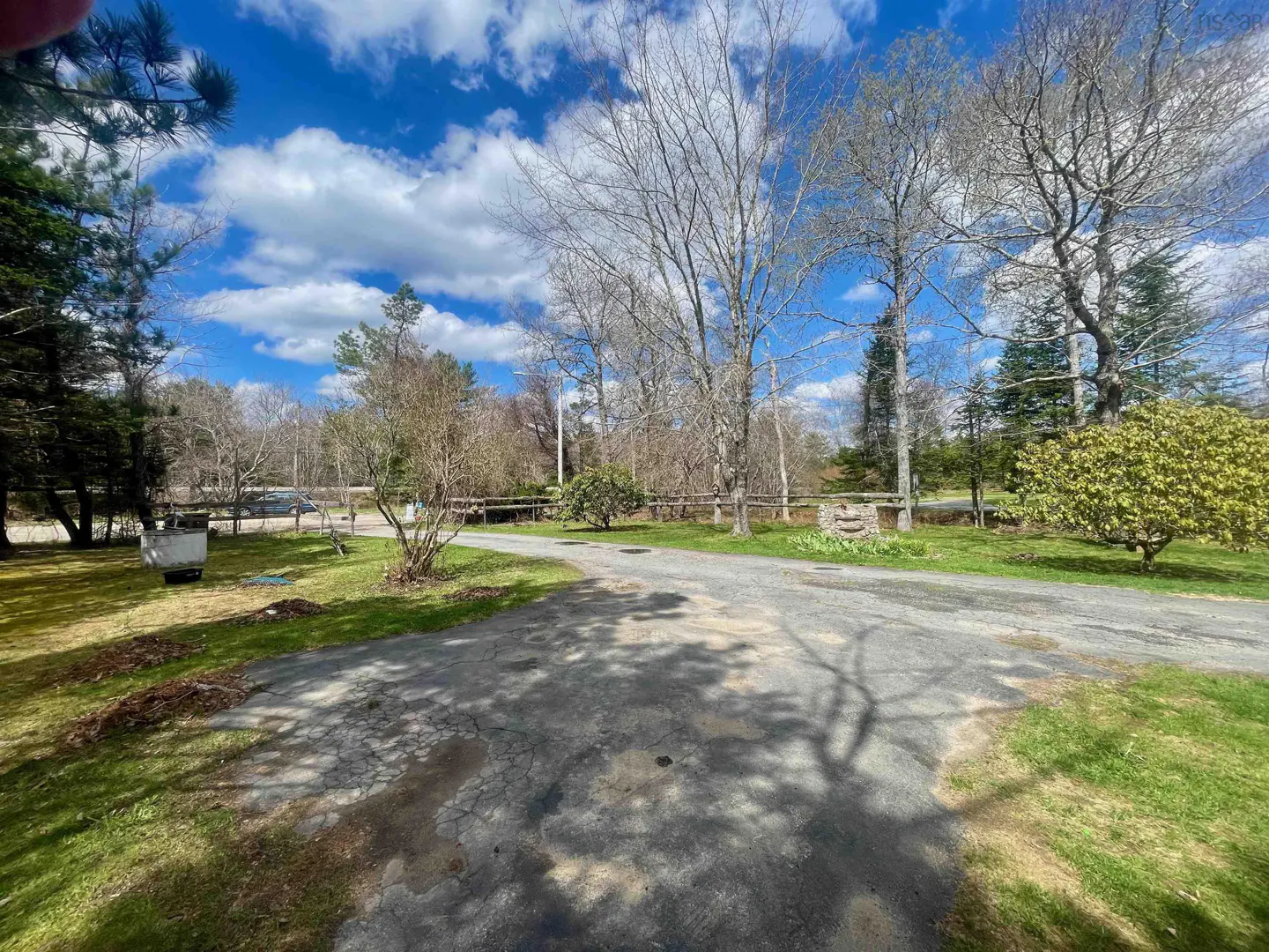 A cracked asphalt driveway leads to a house with trees, grass, and a blue sky with white clouds.