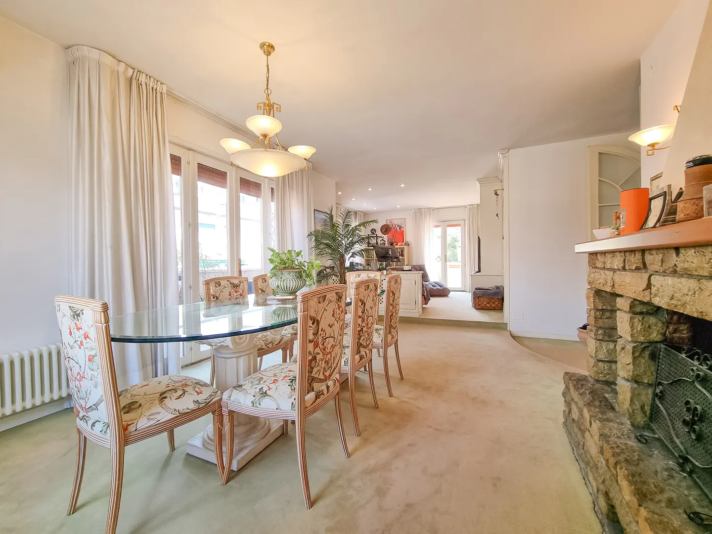 Dining area with glass table, floral chairs, and chandelier, next to a stone fireplace in a spacious, carpeted living room.