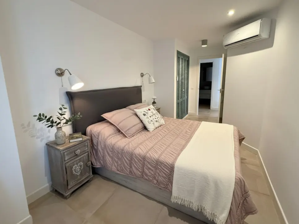 Bedroom with a gray headboard, pink quilt, white throw, and a rustic nightstand with a lamp and greenery.