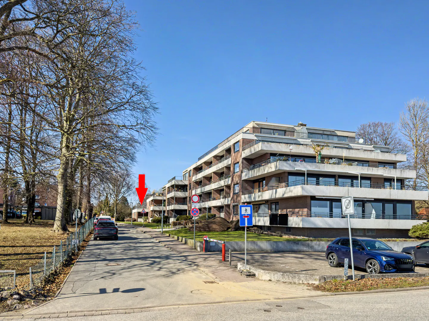 Exterior view of a modern, multi-story apartment building with balconies, cars parked in front, and street signs.