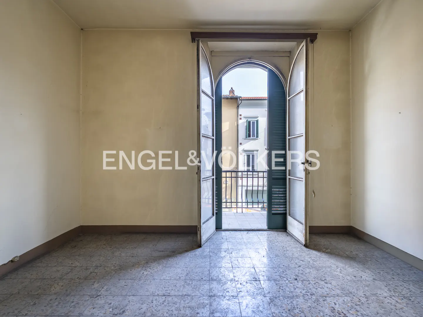 Empty room with beige walls and tiled floor. Open arched doors lead to a balcony with a view of buildings.
