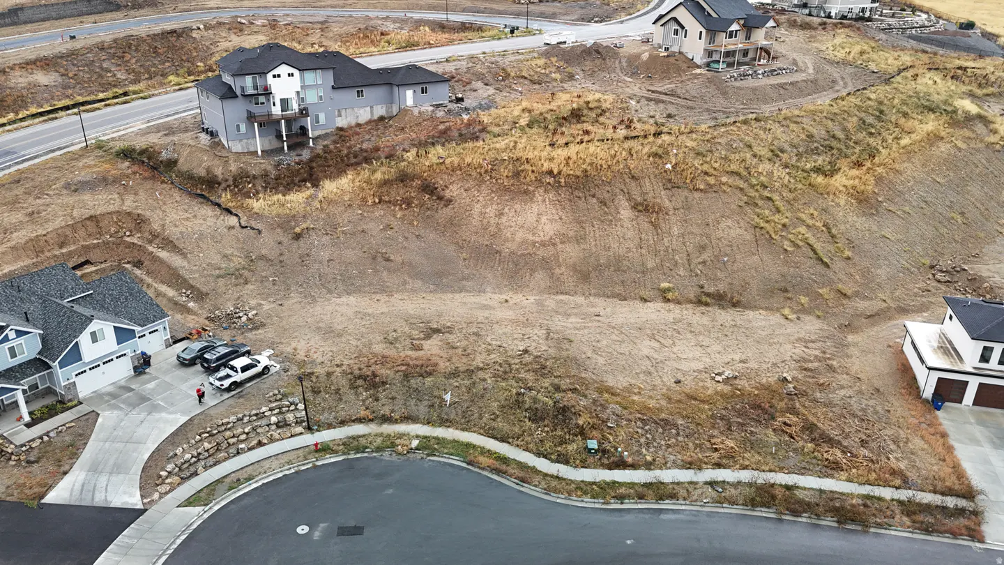 Aerial view of a vacant lot on a hillside in a residential area, with houses nearby. The lot is mostly bare dirt with some dry grass.