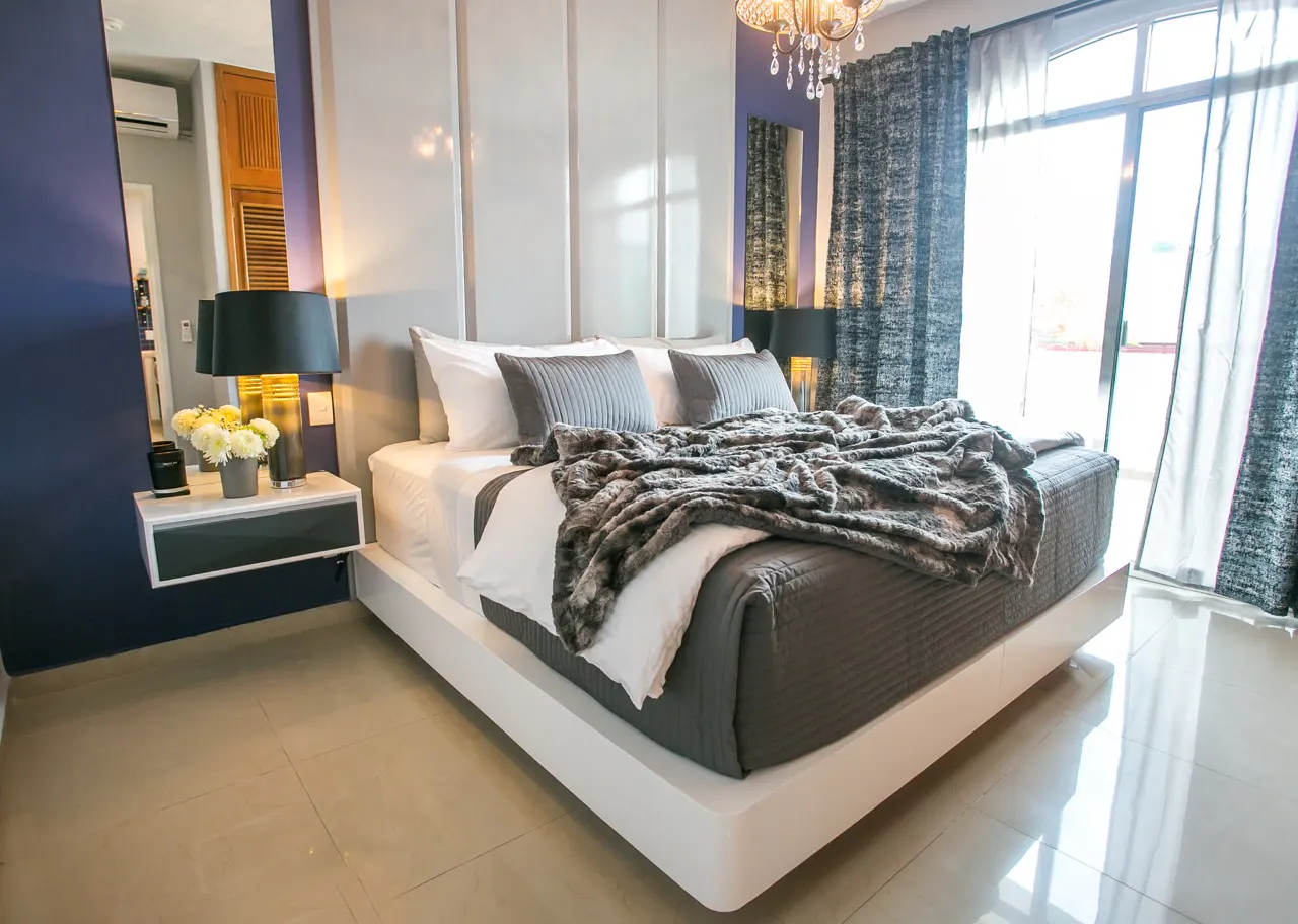 Bedroom with a white platform bed, gray bedding, and a faux fur throw. A nightstand with a lamp and flowers sits beside the bed. Large windows with curtains.