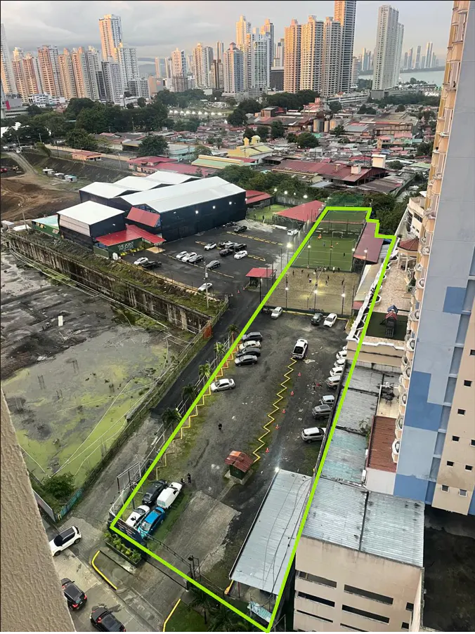 Aerial view of a property outlined in green, featuring parking, tennis courts, and a cityscape backdrop.