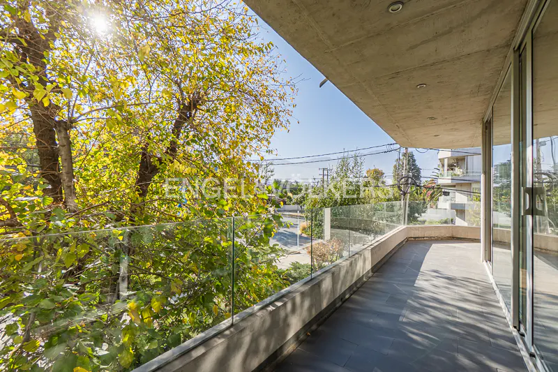 Balcony view with glass railing, gray tile floor, and trees with yellow leaves. Sliding glass doors lead indoors.