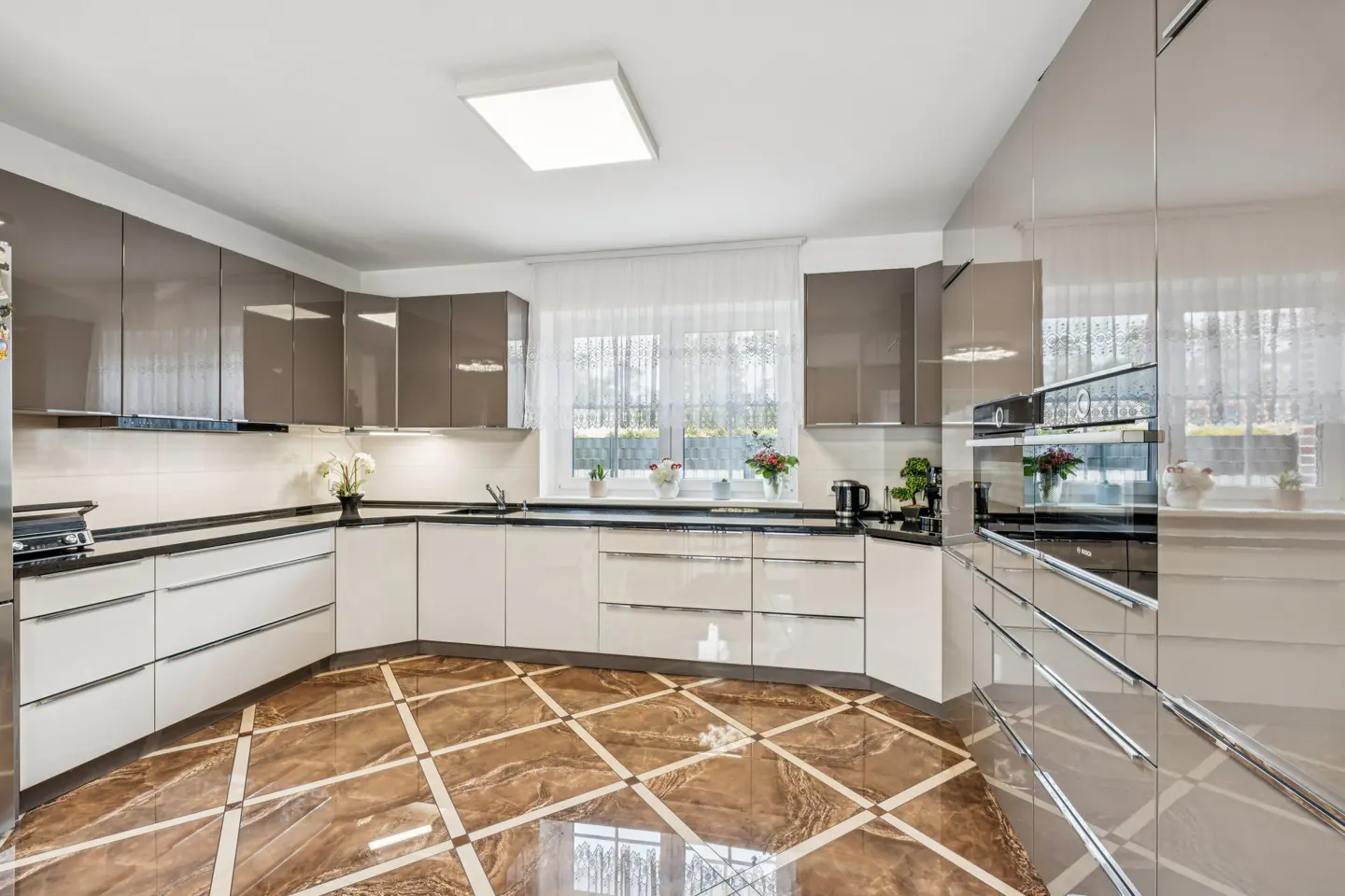 Bright kitchen with white and gray cabinets, black countertops, and brown patterned tile floor. A window with sheer curtains is above the sink.