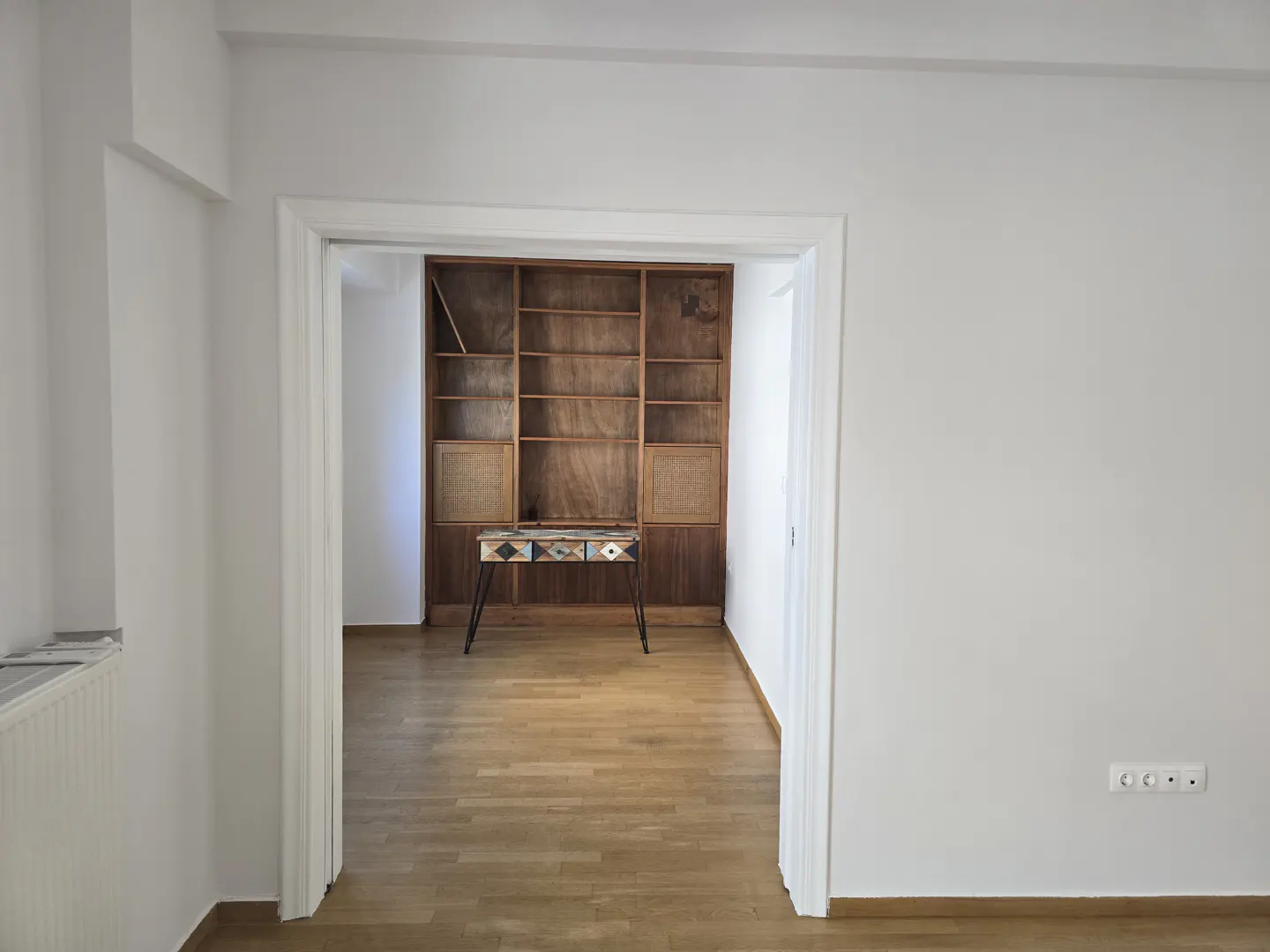 View through a doorway into a room with a wooden bookshelf and a small table on a wood floor in a white-walled room.