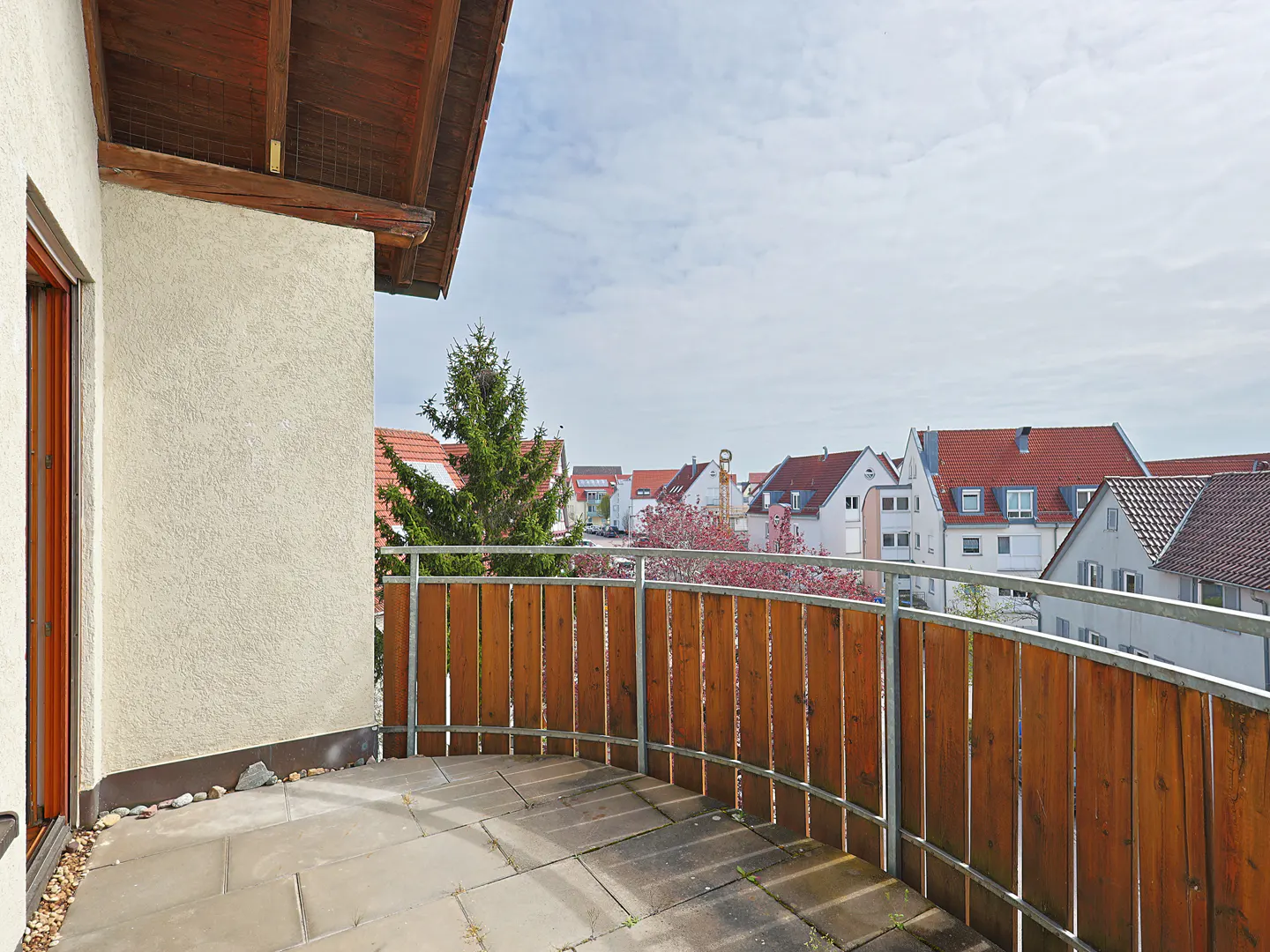 Balcony view with wood and metal railing overlooking houses with red roofs under a cloudy sky.