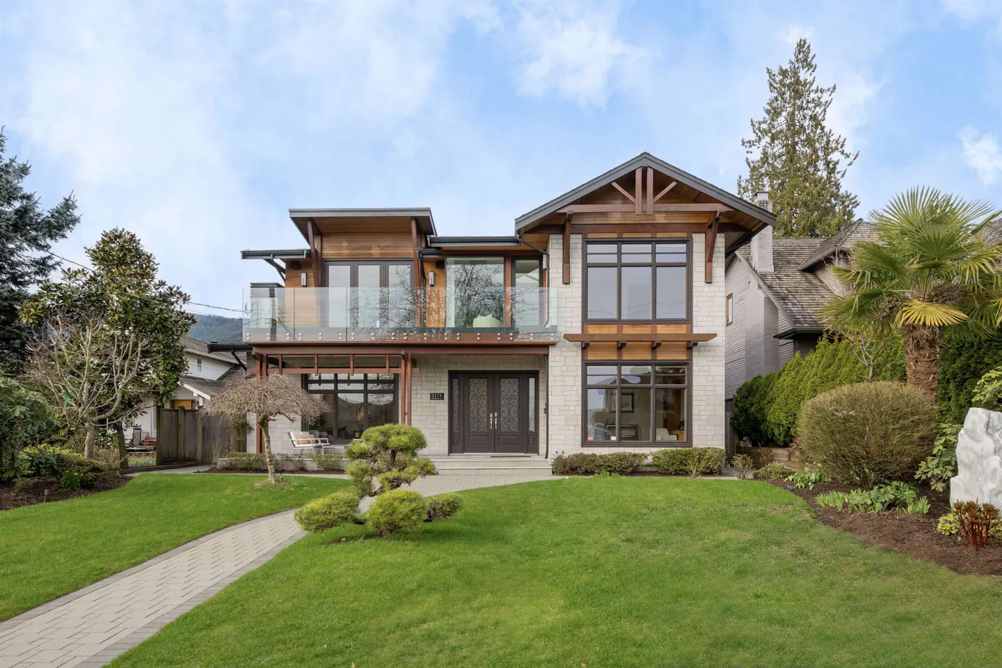 Two-story house with stone and wood exterior, dark trim, and a glass-railed balcony. A green lawn and stone walkway lead to the front door.