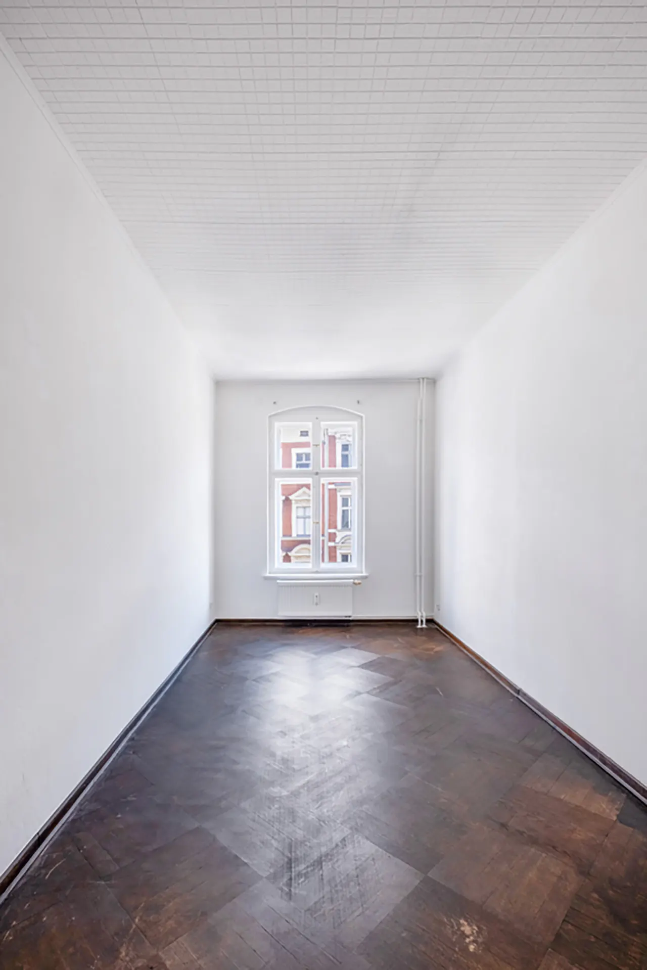 Empty room with white walls, dark parquet floor, and a window showing a red brick building.