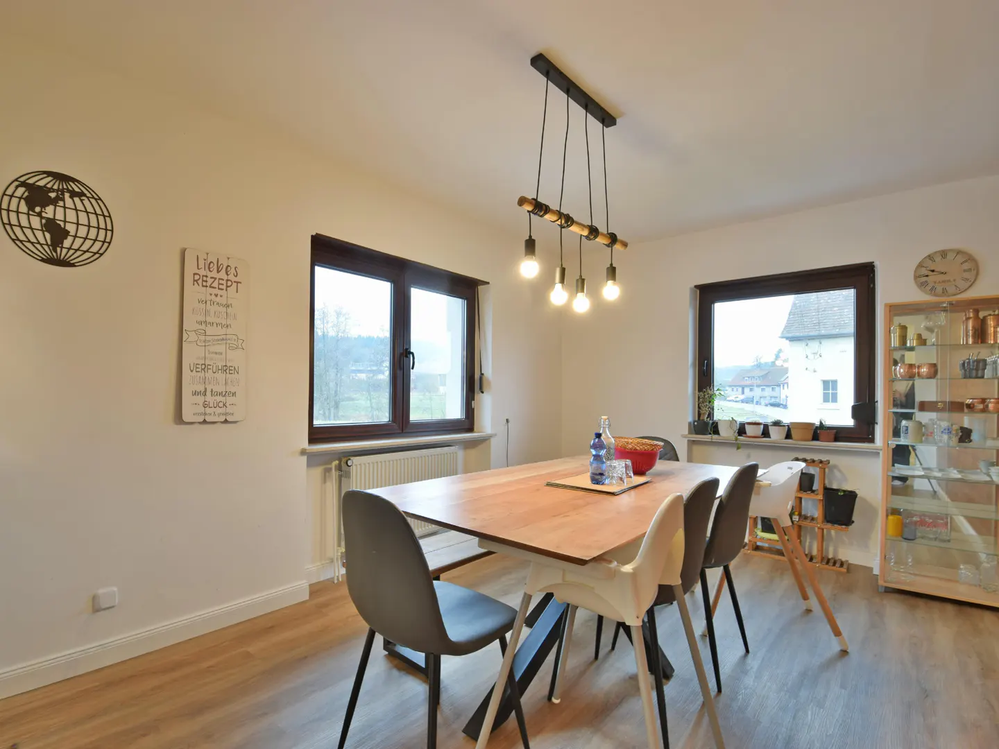 Dining room with wood table, gray chairs, and modern light fixture. Windows show a view of the outside. A glass cabinet is on the right.