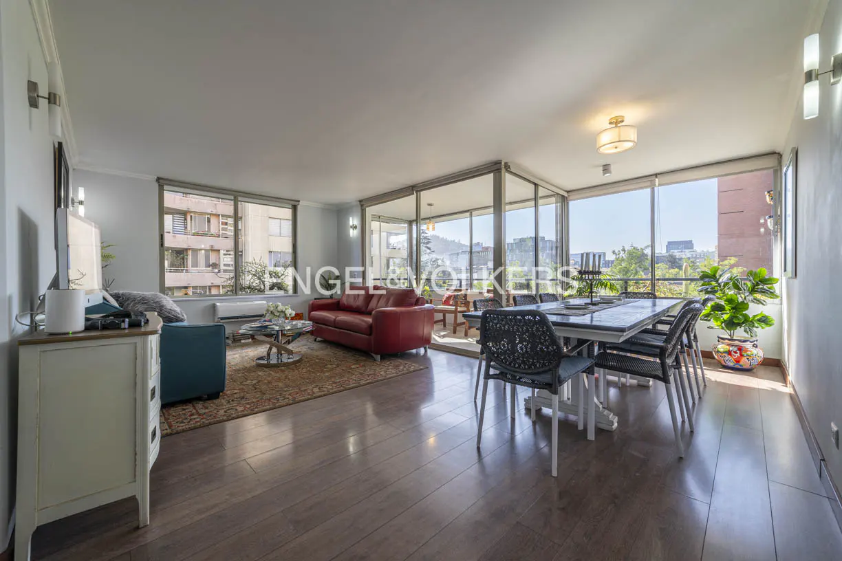 Bright, open-plan living and dining area with wood floors, red sofa, and city views through large windows.