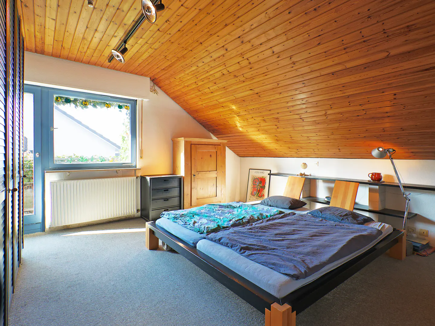Attic bedroom with wood-paneled ceiling, bed with blue covers, window, and built-in shelves.