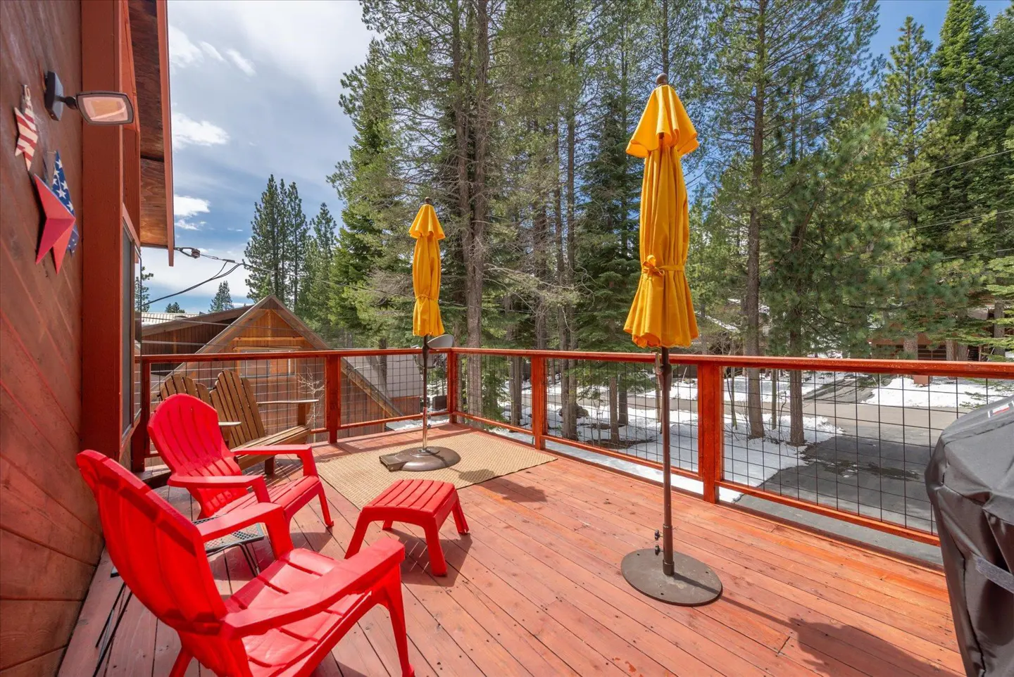 A wooden deck with red Adirondack chairs, yellow umbrellas, and a view of tall trees and snow.