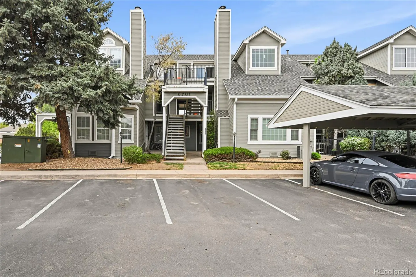 Exterior view of a gray two-story condo building with a gray car parked under a carport.