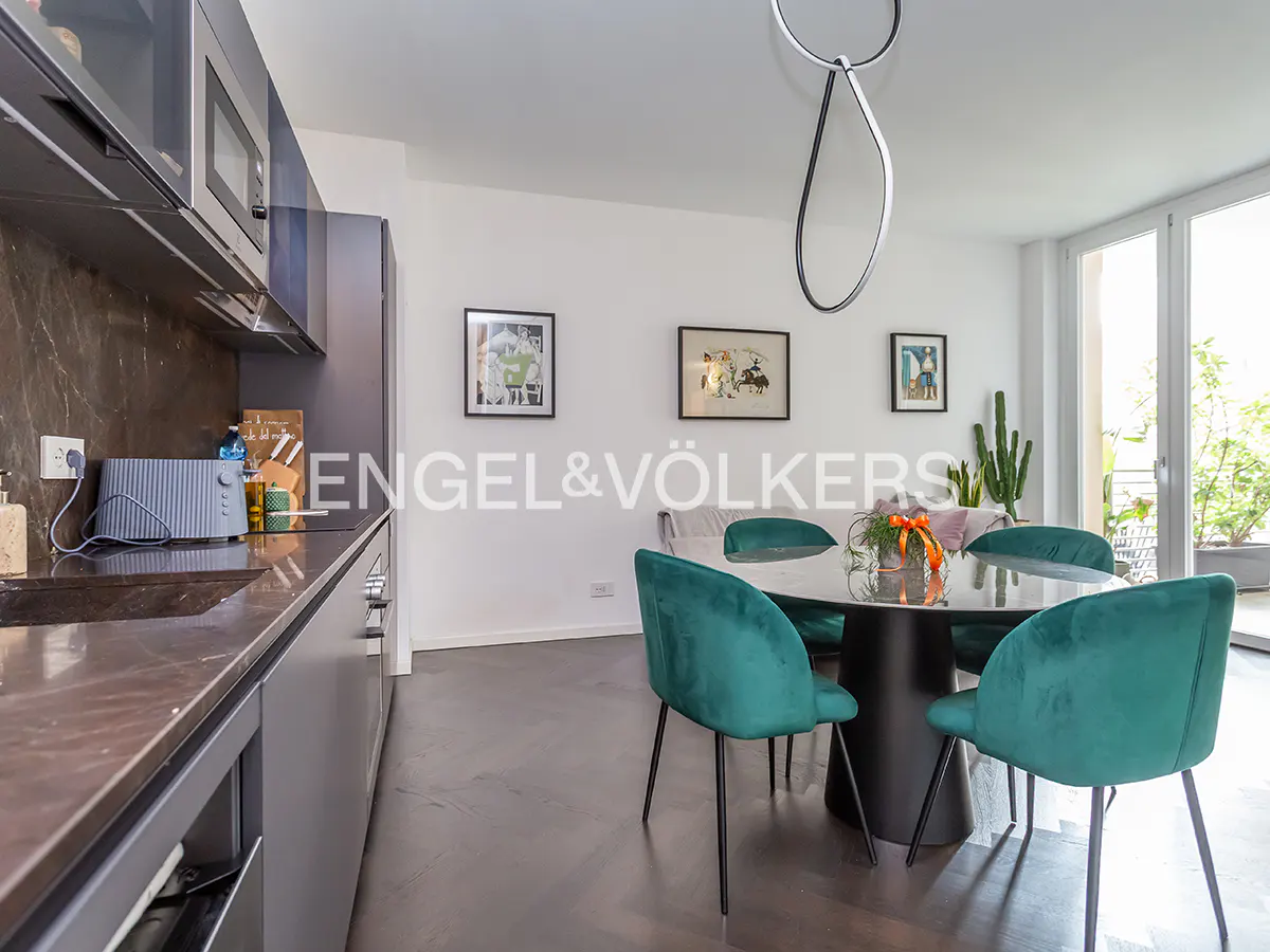 A modern kitchen and dining area with dark cabinets, a marble countertop, and a round table with teal chairs. Artwork adorns the white walls.
