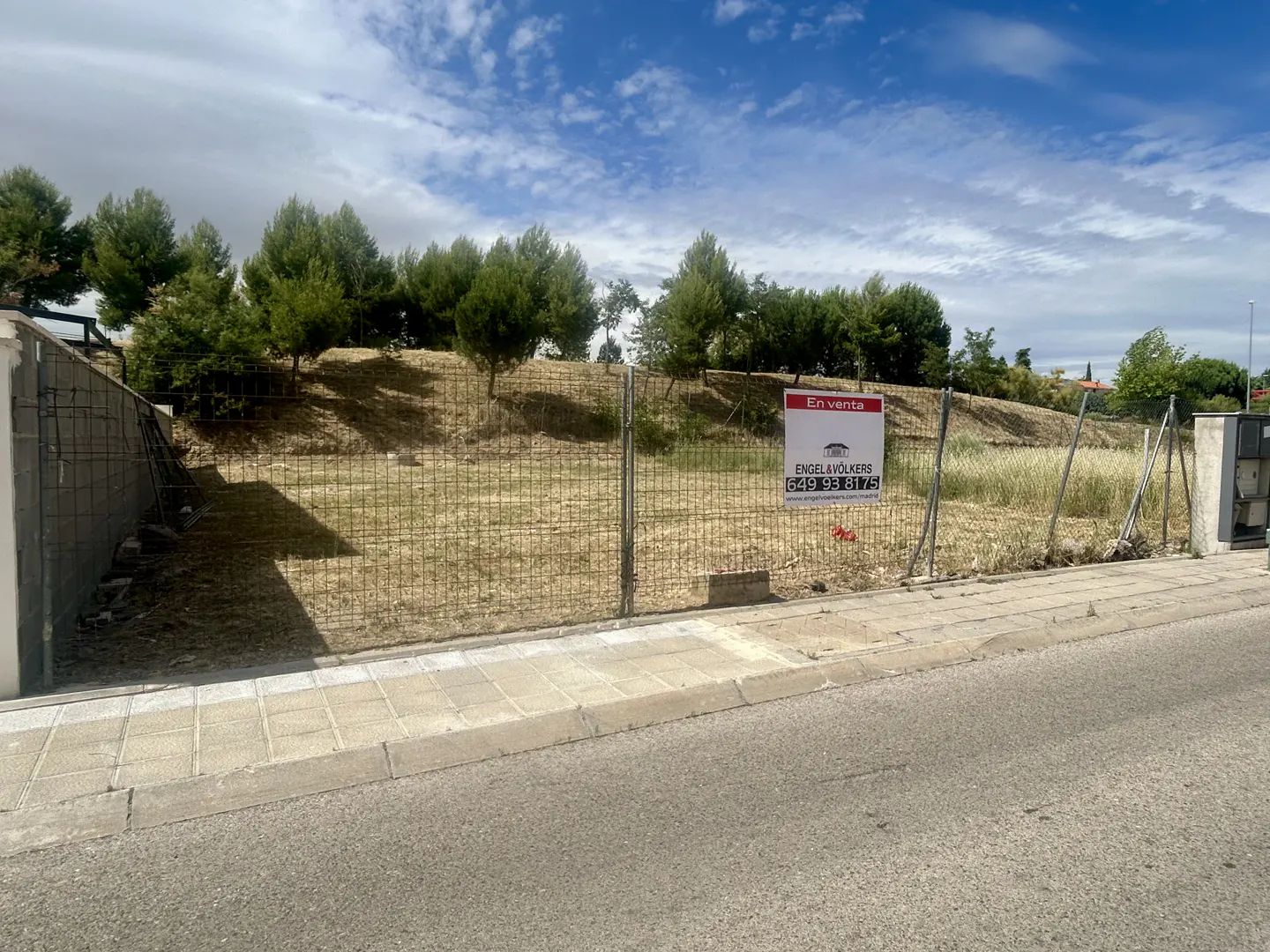 Vacant fenced plot of land for sale with an Engel & Völkers sign, bordered by trees and a sidewalk under a blue sky.