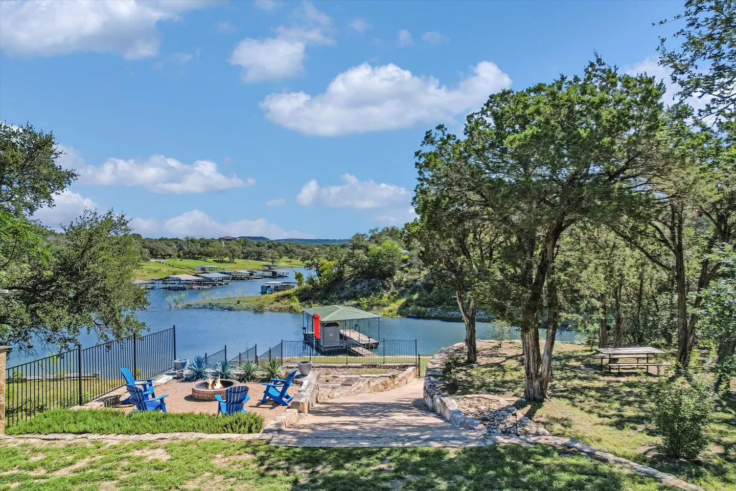 Scenic view of a lake house with blue chairs around a fire pit, a dock, and lush greenery under a blue sky.