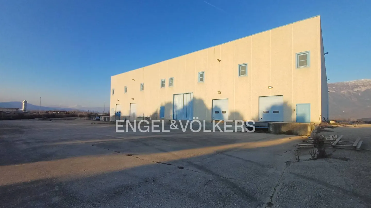Exterior of a large, light yellow warehouse with blue doors and windows under a clear blue sky. Mountains are visible in the background.