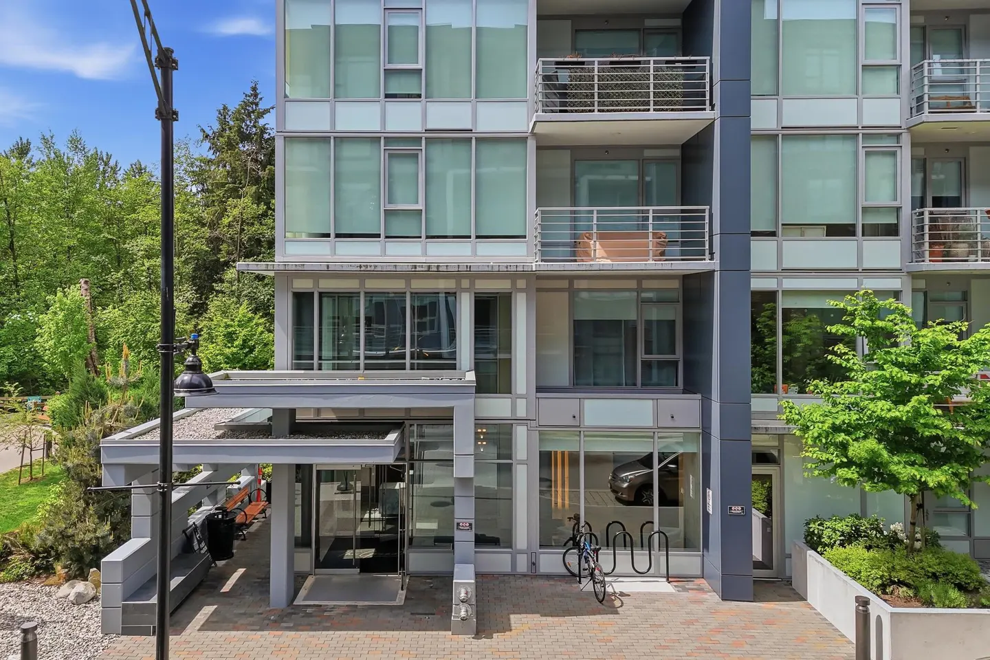Exterior view of a modern, multi-story condo building with glass windows and balconies, surrounded by green trees and a blue sky.