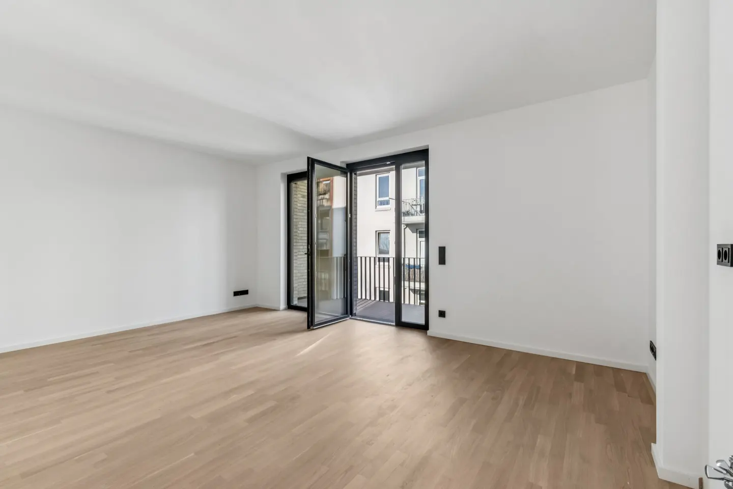 Bright, empty room with light wood floors and white walls. Black framed glass doors open to a balcony with a view of a building.