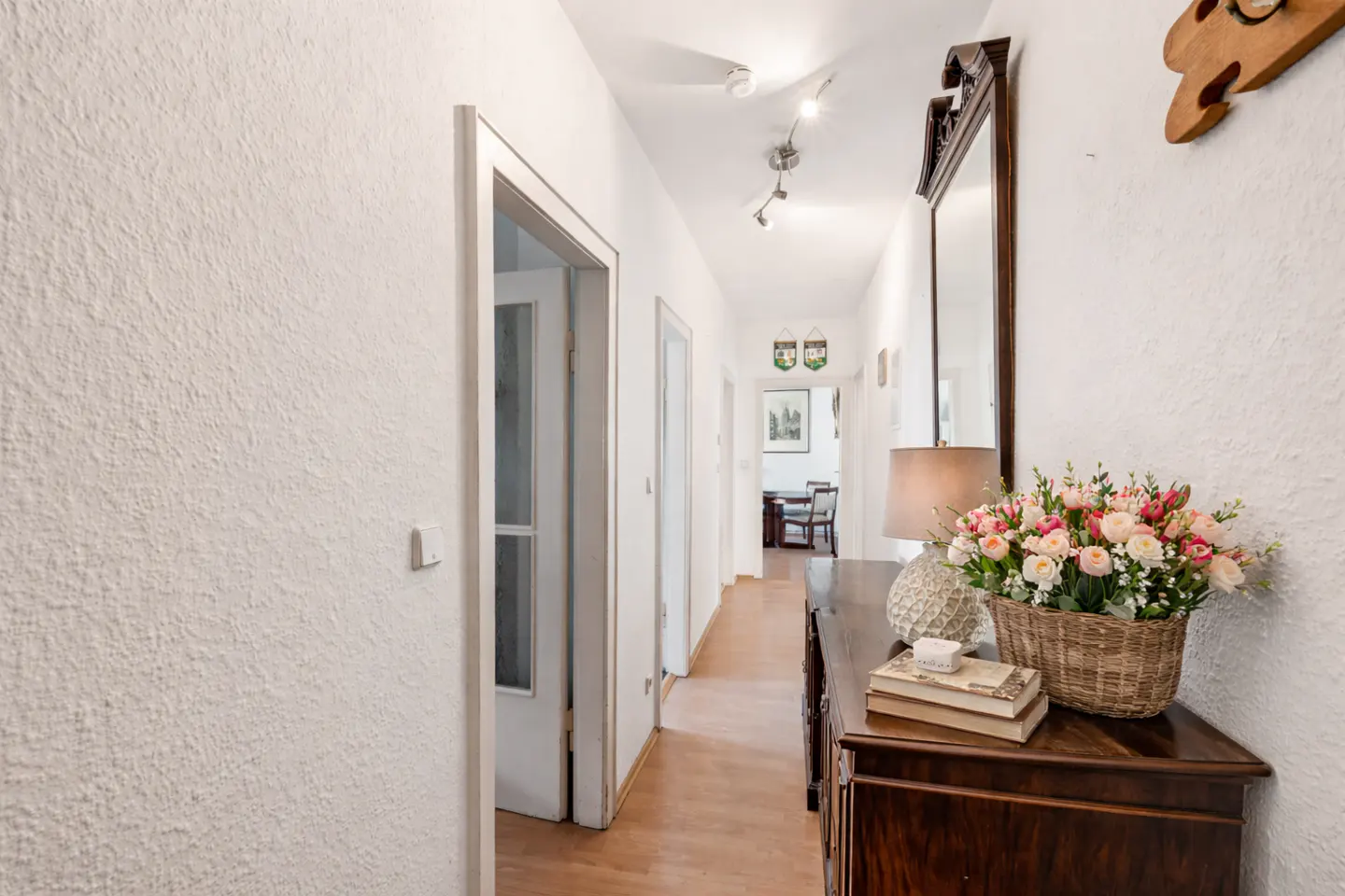 Hallway view with white walls, wood floors, and a dark wood dresser topped with a lamp, books, and a basket of pink and white roses.