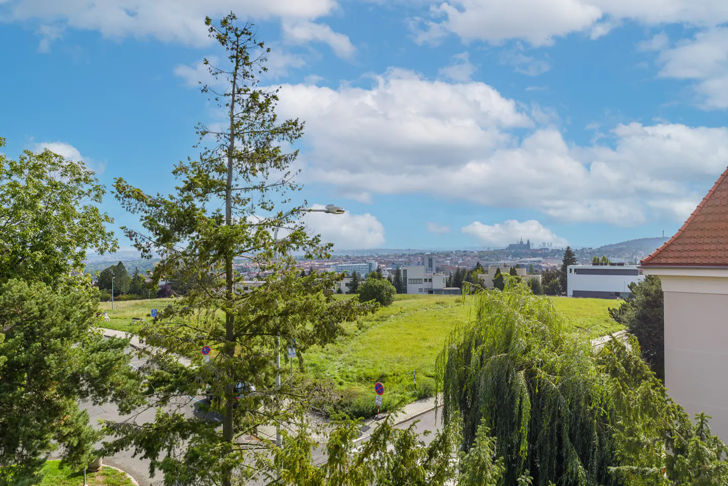 Elevated view of a green field and trees with a distant city skyline under a blue sky with white clouds.
