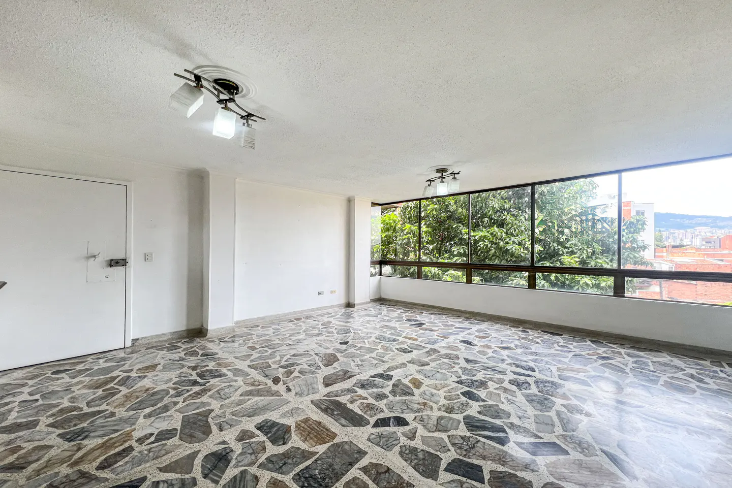 Empty room with white walls, stone tile floor, and large windows showing trees and city view. Ceiling lights are visible.