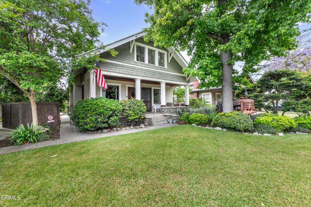 A two-story gray house with a green lawn, bushes, and trees. An American flag hangs on the front porch.