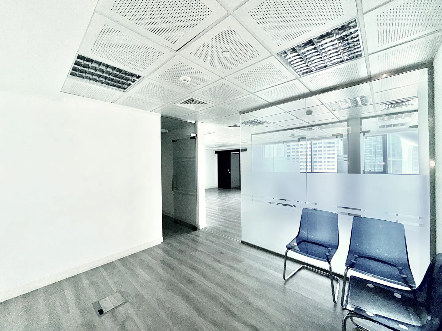Empty office space with gray wood floors, white walls, and a suspended ceiling with fluorescent lights. Blue chairs sit in front of a frosted glass wall.