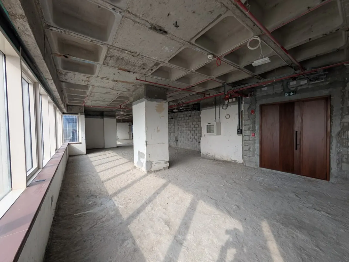 Unfinished office space with concrete floors, walls, and ceiling. Large windows line one side, and a pair of brown doors are on the opposite wall.