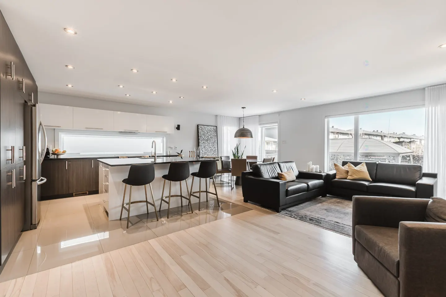 Open-concept living space with kitchen island, black bar stools, dark leather sofas, and light wood floors.